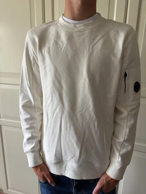 Vit CP Company tröja - Säljer en stilren vit CP Company sweatshirt med rund hals och ribbade muddar. Tröjan har en unik detalj med ficka och svart rund patch på vänster ärm. Perfekt för dig som gillar minimalistisk och clean stil. Tröjan är använd max 3 gånger så väldigt bra skick! Tröjan är i storlek L men passar även M. Hör av er vid frågor och priset är ej hugget i sten!