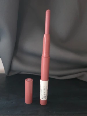 Maybelline SuperStay Matte Ink 15 - Säljer ett Maybelline SuperStay Matte Ink läppstift i färgen 15 Lead The Way. Läppstiftet har en smal, utdragbar form och kommer i en snygg rosa-nude nyans. Perfekt för dig som gillar matta läppar och enkel applicering.