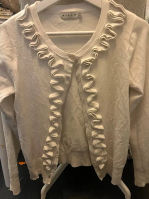 Vit kofta med volang från Micha - Supersöt vit kofta från Micha med dekorativa volanger längs knäppningen och halsringningen. Koftan har långa ärmar och ribbade muddar. Perfekt för dig som gillar en feminin och stilren look.