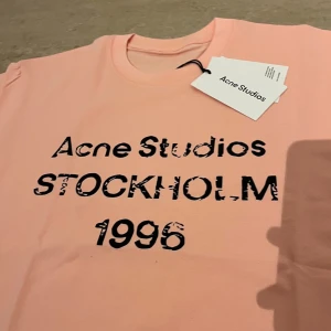 Ljusrosa Acne Studios t-shirt - Säljer en ljusrosa t-shirt från Acne Studios med svart tryck framtill där det står 'Acne Studios STOCKHOLM 1996'. Klassisk rund hals och korta ärmar. Perfekt för dig som gillar stilrena plagg med en twist.