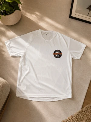 Vit Dolphins basket t-shirt - Vit t-shirt med Norrköping Dolphins Basketball Camp-logga på bröstet. Tillverkad i ett lätt och ventilerande material, perfekt för sport och träning. Klassisk passform med rund hals och korta ärmar. Köpt på en match i Norrköping 
