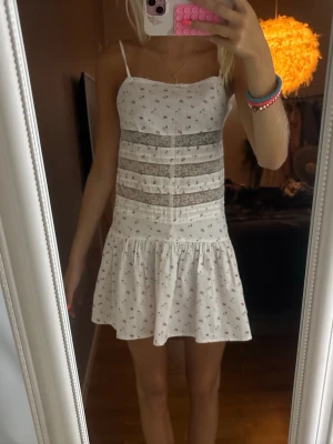 Vit kort sommarklänning med spets - Söt vit kortklänning med smala axelband och volangkant nertill. Klänningen har små blommiga mönster och två breda spetsdetaljer över magen. Aldrig använt 