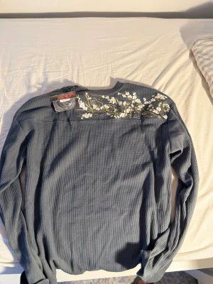 Dershutze waffle longsleave - Mysig blå stickad tröja med lång ärm och rund hals. Tröjan har ett snyggt blommönster i vitt och grönt över axeln och är tillverkad i ett ribbat material som ger en avslappnad look. Perfekt för dig som gillar detaljer och vill ha något unikt i garderoben.