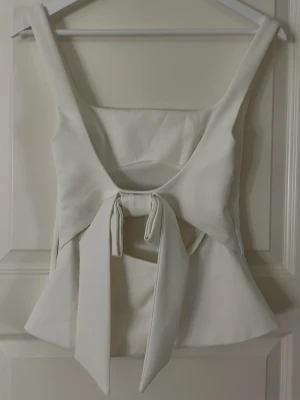 Vit omlott topp med knyt - Snygg vit topp med breda axelband och omlott-design som knyts framtill. Toppen har en lätt utsvängd form och är ärmlös, perfekt för att skapa en chic look. Materialet känns som polyester och ger en slät finish.