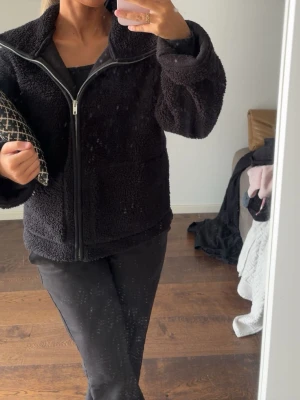 Svart teddyjacka med dragkedja - Mysig svart teddyjacka med hög krage och dragkedja framtill. Jackan har en oversized passform och stora fickor på sidorna. Perfekt för kalla dagar och ger en trendig look med sin fluffiga struktur.
