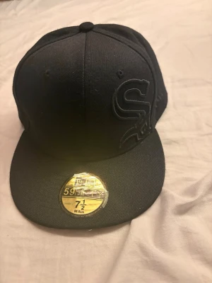 Svart sox keps - Säljer en svart 59FIFTY keps med platt skärm och broderad logga framtill. Klassisk modell med klistermärke kvar på skärmen. Tillverkad i ett tåligt tygmaterial och har ventilerande hål upptill. Perfekt för dig som gillar streetwear och stilrena accessoarer.