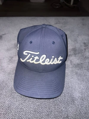 Mörkblå Titleist keps - Snygg mörkblå keps från Titleist med broderad logga framtill i vitt. Klassisk böjd skärm och justerbar passform. Tillverkad i ett slitstarkt material som passar perfekt för golf eller vardagsstil.