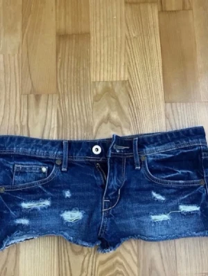 Lågmidjade jeansshorts - Säljer ett par mörkblå jeansshorts med slitna detaljer och råa kanter. Shortsen har klassisk femficksdesign, bälteshällor och dragkedja med knapp. Perfekta för en avslappnad och trendig look.