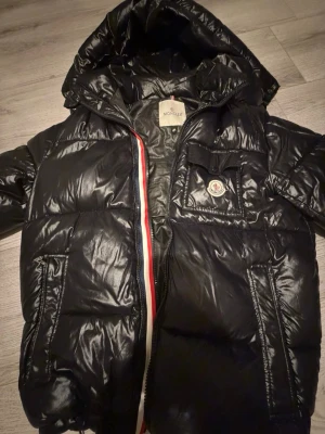 Svart dunjacka från Moncler med huva - Snygg svart dunjacka från Moncler med glansig finish och huva. Jackan har dragkedja med röd, vit och blå detalj, samt en bröstficka med Moncler-logga. Två sidofickor med dragkedja och puffig design som håller dig varm under vintern.