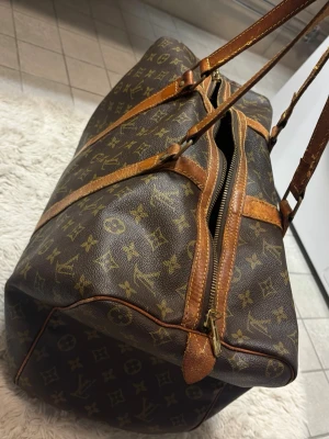 Louis Vuitton Monogram weekendbag - Snygg brun weekendbag från Louis Vuitton med klassiskt monogrammönster och detaljer i läder. Väskan har dubbla handtag, dragkedja och är  rymlig perfekt för weekend bag eller handbagage för resor eller övernattningar.  Lagad i handtaget se bild, minimaliskt hål där bult och läder lapp suttit en gång i tiden, se bild, den andra finns kvar på andra sidan av väskan, slitage framkommer absolut då väskan är från 1987, men desto mer användning desto snyggare om du frågar mig, pris kan diskuteras 