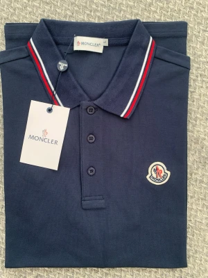 Marinblå pikétröja från Moncler - Snygg marinblå pikétröja från Moncler med klassisk krage och knäppning framtill. Kragen har röda och vita ränder och bröstet pryds av Monclers ikoniska logotyp. Perfekt till jeans eller shorts för en clean look.