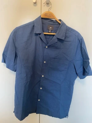 Blå kortärmad skjorta från H&M - Snygg blå skjorta från H&M med korta ärmar och klassisk krage. Skjortan har knappar framtill och en bröstficka. Tillverkad i ett lätt och svalt bomullsmaterial, perfekt för varma dagar.