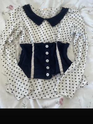 Prickig blus med marinblå detaljer - Söt vit blus med svarta prickar, långärmad och med marinblå krage. Midjan har en marinblå sektion med vita knappar och spetsdetaljer. Blusen har en feminin och retroinspirerad look, perfekt för dig som gillar unika plagg.