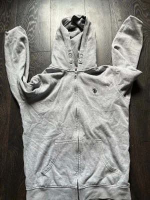 Grå zip hoodie med broderad logga - Säljer en grå hoodie med dragkedja och huva. Tröjan har en broderad logga på bröstet, kängurufickor och ribbade muddar. Perfekt för chill dagar eller när du vill ha en enkel och clean look.