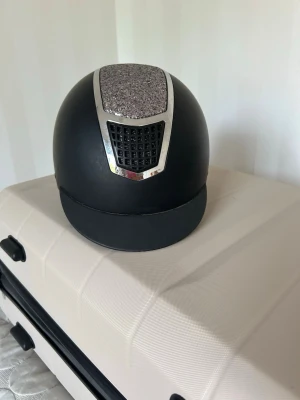 Svart ridhjälm med glitterdetalj - Snygg svart ridhjälm med en stor glittrig detalj på ovansidan och silvriga ventiler framtill och bak. Hjälmen har en klassisk rund form och skärm framtill. Materialet är matt och ger ett stilrent intryck, perfekt för dig som vill sticka ut i stallet.