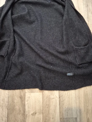 Mörkgrå kofta med fickor - Mysig mörkgrå kofta i mjukt material med två stora fickor framtill. Koftan har en avslappnad passform och ribbade muddar nedtill. Perfekt att slänga över en t-shirt eller topp för extra värme.