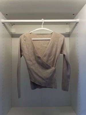 Beige omlottopp med ribbad struktur - Snygg beige omlottopp med ribbad struktur och lång ärm. Toppen har en djup v-ringning och sitter tight på kroppen. Perfekt för dig som gillar en croppad och stilren look.