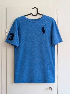 Polo Ralph Lauren Big Pony T-shirt blå – XL 18–20 - Snygg blå T-shirt från Polo Ralph Lauren med stor broderad Big Pony-logga på bröstet och klassisk 3-detalj på ärmen. Blåmelerat tyg med mörkblå detaljer ger en sportig/preppy look. 👕 Storlek: XL (18–20 år) motsvarar ca xs/s i vuxenstorlek! 🎨 Färg: blåmelerad / mörkblå detaljer ✨ Skick: Bra men den är lite nopprig! 📏 Mått kan skickas vid intresse 📦 Skickas snabbt