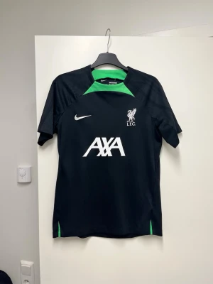 Svart Nike Liverpool träningströja XL - Säljer en svart träningströja från Nike med gröna detaljer och Liverpool FC-logga på bröstet. Tröjan har Dri-Fit-material som andas och är i slim fit-modell. AXA-logga framtill och klassisk Nike-swoosh. Perfekt för fotbollsträning eller gym.