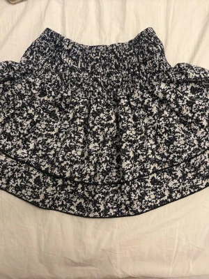 Svartvit blommig kortkjol - Supersöt kortkjol med svartvitt blommönster och bred resår i midjan. Kjolen har en luftig och volangig form som ger en avslappnad vibe. Perfekt för dig som gillar en feminin och trendig stil.
