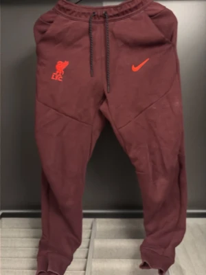 Vinröda Nike Liverpool mjukisbyxor - Säljer ett par vinröda mjukisbyxor från Nike med Liverpool FC-logga och Nike Swoosh i rött framtill. Byxorna har dragsko i midjan, muddar vid bensluten och är tillverkade i mjukt material. Perfekta för chill dagar eller när du vill representera ditt lag. Går att köpa till tröjan för 150 men den är lite sönder vid fickorna