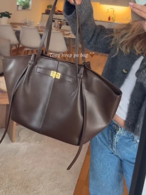 Mörkbrun stor handväska med lång rem - Snygg mörkbrun handväska i PU-läder med lång axelrem och guldfärgat spänne framtill. Väskan har en rymlig design med vingsidor och minimalistiska sömmar. Perfekt för dig som vill ha plats för allt du behöver under dagen. Ny med prislapp 