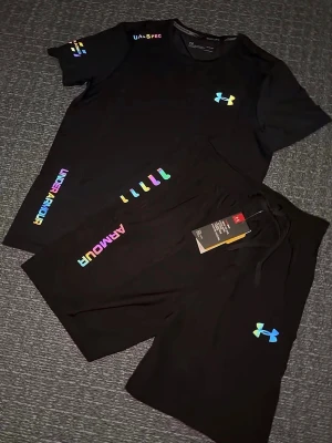 Svart träningsset från Under Armour  - Säljer ett par svart träningsset från Under Armour med regnbågsfärgade loggor och textdetaljer. Shortsen har snörning i midjan och är perfekta för träning eller avslappnade dagar. Matchande t-shirt finns också med samma färgglada detaljer. Storlek S, helt ny 