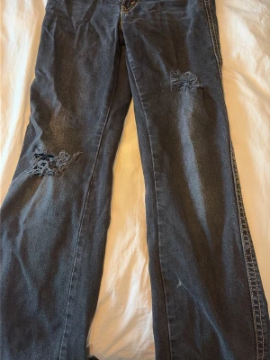 Svarta jeans med utsvängda ben och hål vid knäna  - Säljer ett par svarta lågmidjade  jeans med utsvängda ben och hål på båda knäna. Jeansen har klassisk femficksdesign och kontrastsömmar längs sidorna.