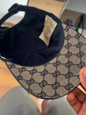 Gucci beige keps med GG-mönster - Säljer en beige keps från Gucci med klassiskt GG-monogram i mörkblått över hela kepsen. Justerbar rem bak och en snygg detalj med röd och marinblå rand. Tillverkad i canvasmaterial med mörkblå insida. Perfekt för dig som gillar exklusiva accessoarer.