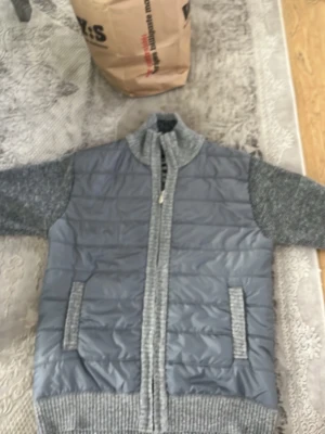 Grå quiltad cardigan med stickade ärmar - Snygg grå cardiganjacka med quiltad framsida och stickade ärmar samt rygg. Dragkedja framtill, två fickor och ribbade muddar. Perfekt för lager på lager och chill stil. Materialmix av vadderad polyester och mjukt stickat tyg.