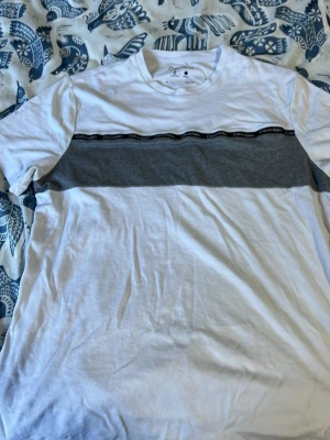 Vit och grå t-shirt från Calvin Klein - Snygg vit t-shirt från Calvin Klein med en bred grå panel över bröstet och svart band med logga. Klassisk rund hals och korta ärmar. Perfekt till jeans eller shorts för en clean och modern look.