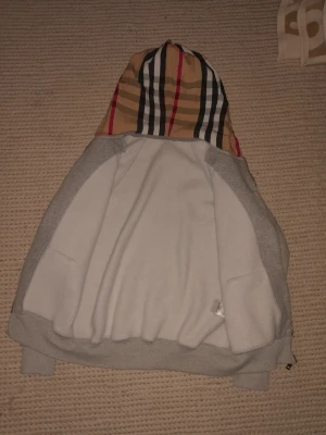Burberry hoodie - Snygg hoodie i ljusgrått med en huva i klassiskt rutigt mönster i beige, svart, vitt och rött. Hoodien har ribbade muddar vid ärmslut och nederkant. Perfekt för en avslappnad och trendig stil.