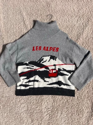 Grå stickad tröja med Les Alpes-motiv - Super skön stickad tröja, med motiv av Alperna. Tröjan är i grått med hög krage och texten 'LES ALPES' i rött. Tröjan är i strl XL men skuller mer säga S/M