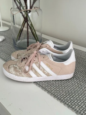 Adidas Gazelle beige spets sneakers - Snygga Adidas Gazelle sneakers i beige mocka med dit satta vita detaljer och spetsmönster på ovandelen. Är hemma gjord och har inga defekter på skorna mer än den tillsatta spetsen. Klassiska tre ränder på sidan och vit sula. Skorna har rosa skosnören och gulddetaljer på sidan och plösen. Perfekta för dig som vill ha en unik touch på dina sneakers. Spetsen som är tillsatt har hålt mycket bra och finns inga täcken på slitage, men dem är hemma gjorde så man har det i å tanke.