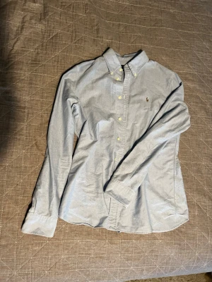 Ljusblå skjorta från Ralph Lauren - Snygg ljusblå skjorta från Ralph Lauren med klassisk krage och knappar framtill. Skjortan har lång ärm och broderad logga på bröstet. Custom fit-modell som sitter riktigt bra och passar till många olika stilar.