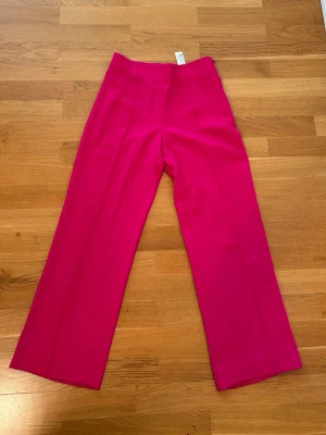Vida kostymbyxor i stark rosa - Säljer ett par vida kostymbyxor i en riktigt stark rosa färg. Byxorna har hög midja och pressveck framtill för en clean look. Perfekta för dig som vill sticka ut och addera färg till din outfit.