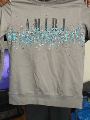 Grå sweatshirt från Amiri med tryck - Säljer en grå sweatshirt från Amiri med stort tryck på bröstet och coolt stänkmönster i blått och svart. Tröjan har rund hals och långa ärmar. Perfekt för dig som gillar streetwear och vill sticka ut lite extra. XL i barn storlek!