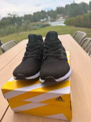 Adidas Ultra Boost svarta sneakers - Svarta Adidas Ultra Boost sneakers med stickad ovandel i mesh för extra andningsförmåga. Skorna har vita Boost-sulor och svarta detaljer med klassiska tre ränder på sidan. Låg modell med snörning och bekväm passform. Perfekta för dig som gillar stilrena och sportiga sneakers.