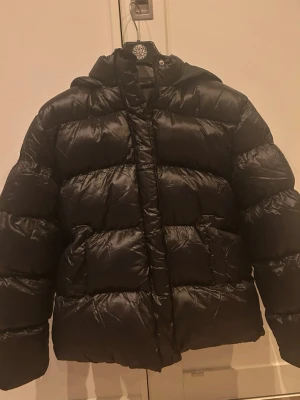 Svart pufferjacka med huva, strl S - Svart pufferjacka i glansigt material med huva och praktiska framfickor. Jackan har dragkedja framtill och en quiltad look som ger extra volym. Perfekt för dig som vill ha en varm och snygg jacka med modern vibe. Jackan är helt ny och aldrig använd. Den är köpt från Novant men det finns ingen lapp på märket i jackan, men jag kan skicka bild på bevis att den är från Novant om intresserad!💕