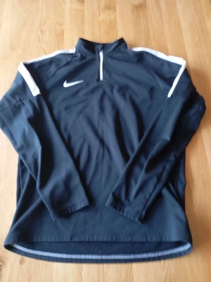 Svart Nike tröja - Svart långärmad träningströja från Nike med vit logga på bröstet och vita detaljer längs axlarna. Tröjan har en halv dragkedja framtill och är tillverkad i ett lätt och funktionellt material som passar perfekt för sport och träning.