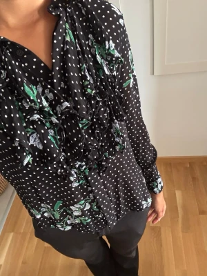 Svart blus med prickar och volanger - Snygg svart blus med vita prickar och grönt blommönster. Blusen har volangdetaljer framtill och knäppning vid halsen. Långärmad modell i ett tunt, mjukt tyg som ger en skön känsla. Perfekt för dig som vill ha något unikt och trendigt.