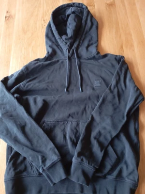Svart boss hoodie - Säljer en svart hoodie från BOSS med klassisk huva och dragsnören. På bröstet sitter en snygg BOSS-logga i tygpatch. Hoodien har känguruficka framtill och ribbade muddar vid ärmar och nederkant. Perfekt för en chill och stilren look.
