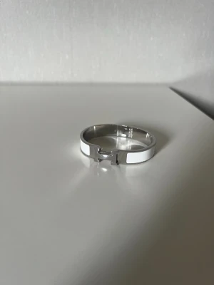 H armband vit/silverfärgat  - Stilrent armband i silverfärgad metall med en bred, slät design och ett diskret H spänne framtill. Perfekt för dig som gillar minimalistiska accessoarer med modern känsla. Passar till många olika stilar och är lätt att matcha med andra smycken.