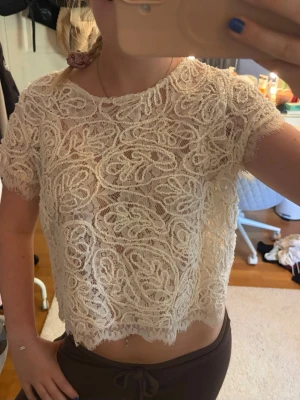 Beige spetsblus med dragkedja - Säljer en beige, croppad spetsblus med korta ärmar och dekorativt mönster. Toppen har en synlig dragkedja i ryggen och vågig kant nertill. Perfekt för dig som gillar romantisk och trendig stil. Skriv pm vid funderingar!☺️