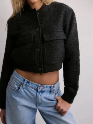 Svart cropped cardiganjacka Nelly - Trendig svart cardiganjacka från Nelly i croppad modell. Jackan har långa ärmar, rund halsringning och stora bröstfickor med lock. Stängs med knappar framtill och har ribbade muddar vid ärmslut och nederkant. Materialet är strukturerat och ger en cool look. Med prislapp