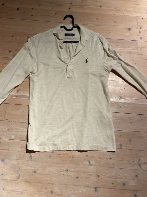 Beige långärmad topp Polo Ralph Lauren - Klassisk beige långärmad topp från Polo Ralph Lauren med krage och knappar framtill. Diskret logga broderad på bröstet. Tillverkad i mjuk bomull som känns skön mot huden. Perfekt för dig som gillar stilrena och tidlösa plagg.