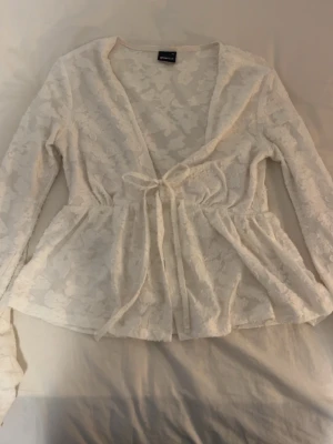 Vit spetskofta från Gina Tricot - Säljer en söt vit spetskofta från Gina Tricot i storlek XS. Koftan har långa ärmar, knytband i midjan och ett romantiskt blommigt spetsmönster. Perfekt att slänga över ett linne eller klänning för en extra touch.