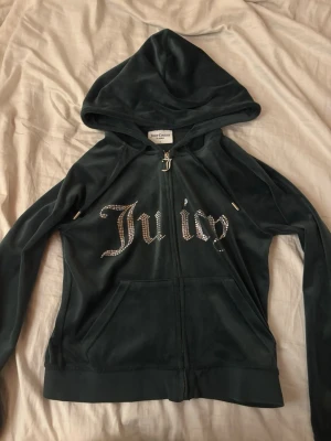 Juicy Couture hoodie  - Mörkgrön hoodie i mjuk sammet från Juicy Couture med glittrande stenar som bildar texten 'Juicy' på framsidan. Dragkedja med J-berlock och två fickor framtill. Perfekt för en trendig och avslappnad look.