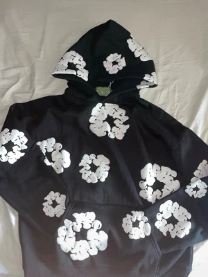 Svart hoodie med vita grafiska blommor - Snygg svart hoodie med stora vita grafiska blommor tryckta över hela plagget. Hoodien har en klassisk känguruficka framtill och en rymlig huva. Perfekt för dig som vill sticka ut med en unik och trendig stil.