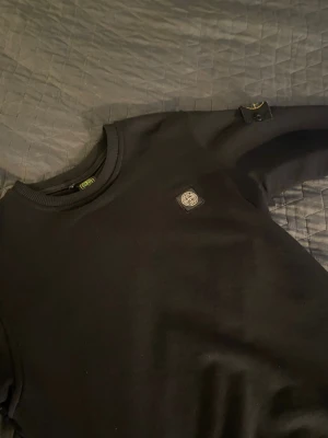 Stone island - Snygg svart stone island tröja med rund hals och ribbade muddar. Tröjan har en liten patch med logga på bröstet och en avtagbar patch på ena ärmen. Perfekt för dig som gillar stilrena och klassiska plagg samt vara den bästa ultran på läktarna